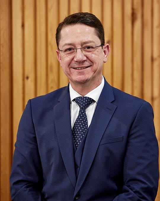 Mann in blauem Anzug und Krawatte, mit Brille, sitzt vor einem Hintergrund aus Holzpaneelen. Dirk Hamacher, Geschäftsführung LACET Niederrhein