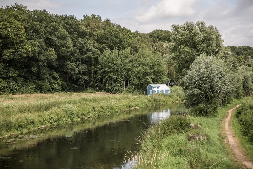 Ein blaues Haus liegt an einem ruhigen Fluss, umgeben von üppigen grünen Bäumen und Feldern, unter einem leicht bewölkten Himmel. Ein schmaler Pfad verläuft am Fluss entlang. Ein blaues Haus liegt an einem ruhigen Fluss, umgeben von üppigen grünen Bäumen und Feldern, unter einem leicht bewölkten Himmel. Ein schmaler Pfad verläuft am Fluss entlang.