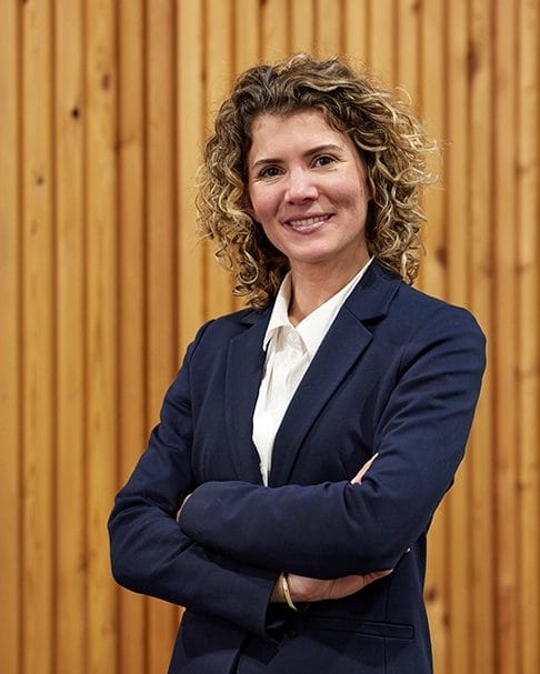 Eine Frau mit lockigem Haar, einem marineblauen Blazer und einem weißen Hemd steht mit verschränkten Armen lächelnd vor einer Holzpaneelwand. Martine Lacet, Management & Prokuristin bei LACET Niederrhein