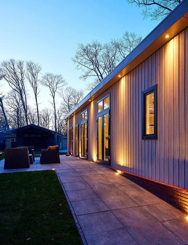 Modernes einstöckiges Gebäude mit Holzverkleidung, großen Fenstern und warmer Außenbeleuchtung in der Dämmerung; Sitzbereich im Freien und blattlose Bäume im Hintergrund.